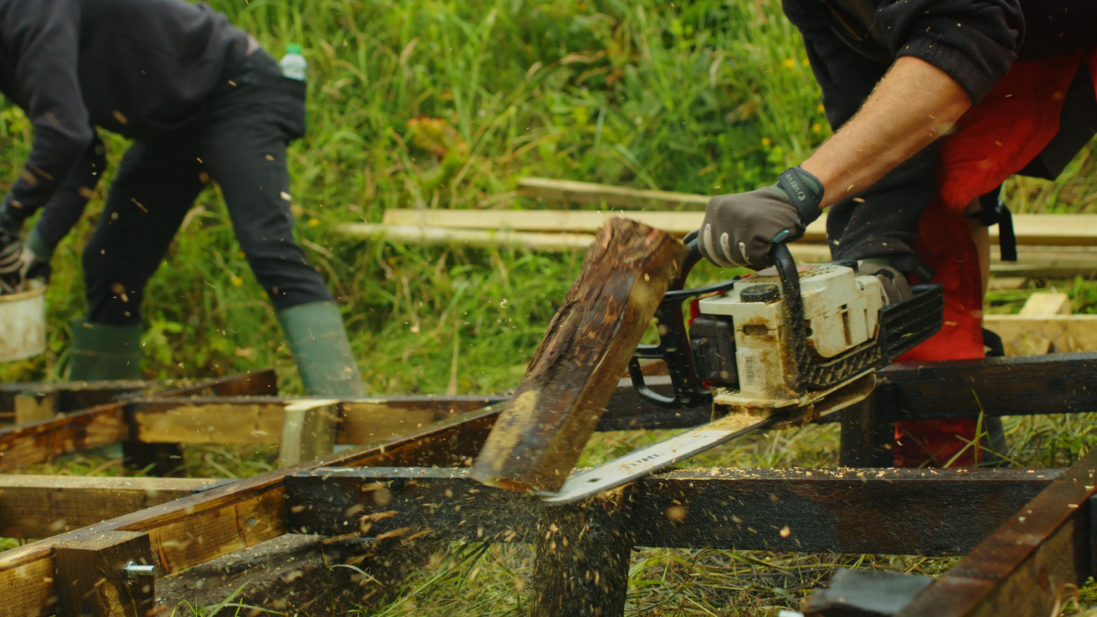 A person uses a chainsaw to cut wood on a grassy field. Another person works nearby, focused. The scene conveys industrious teamwork and focus.